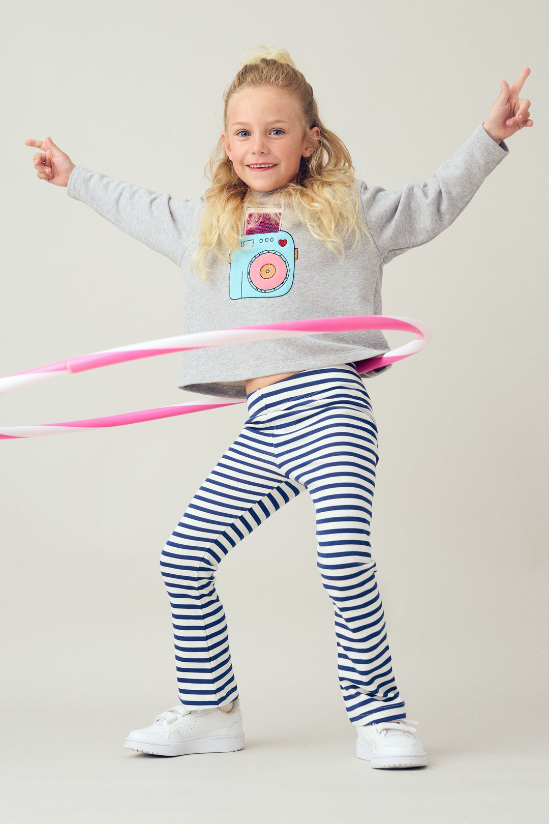 Child wearing a gray sweatshirt with a camera design and striped pants, holding a pink hula hoop.